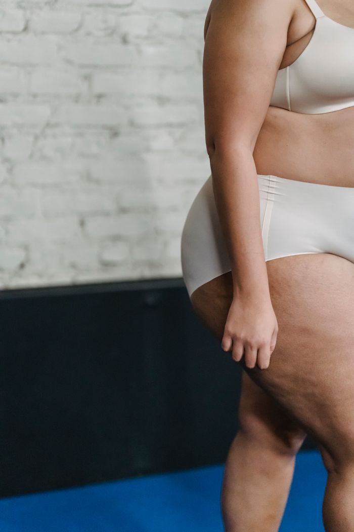 Side view of plus size female in white panties and bra standing on mat near brick wall in light gym