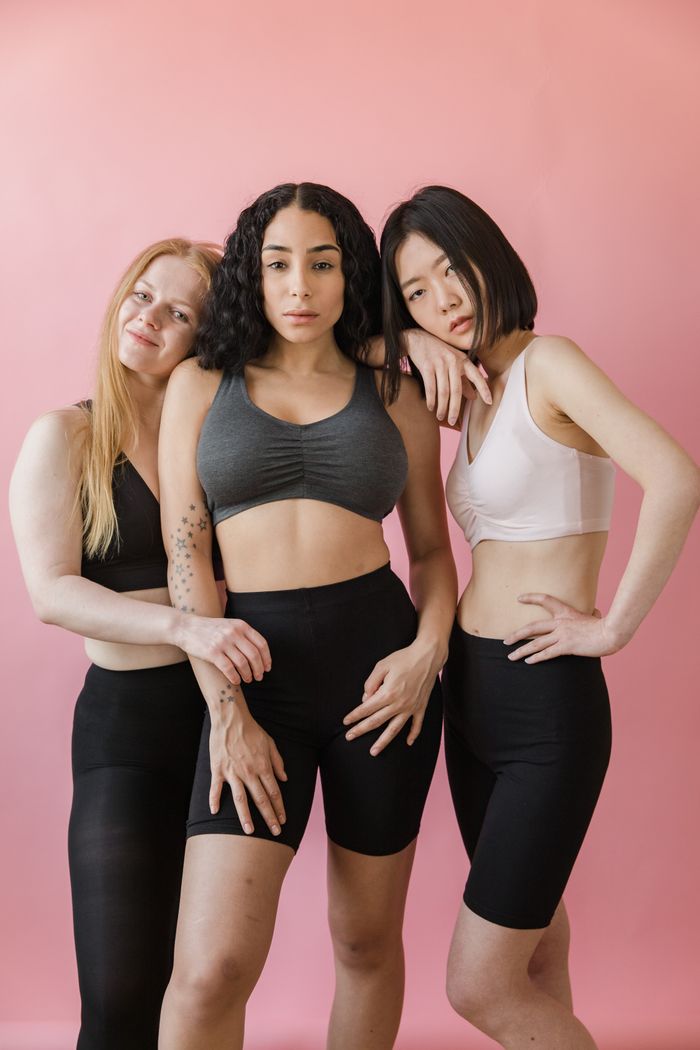 Three diverse women in sports attire confidently stand together against a pink background, embodying strength and unity.